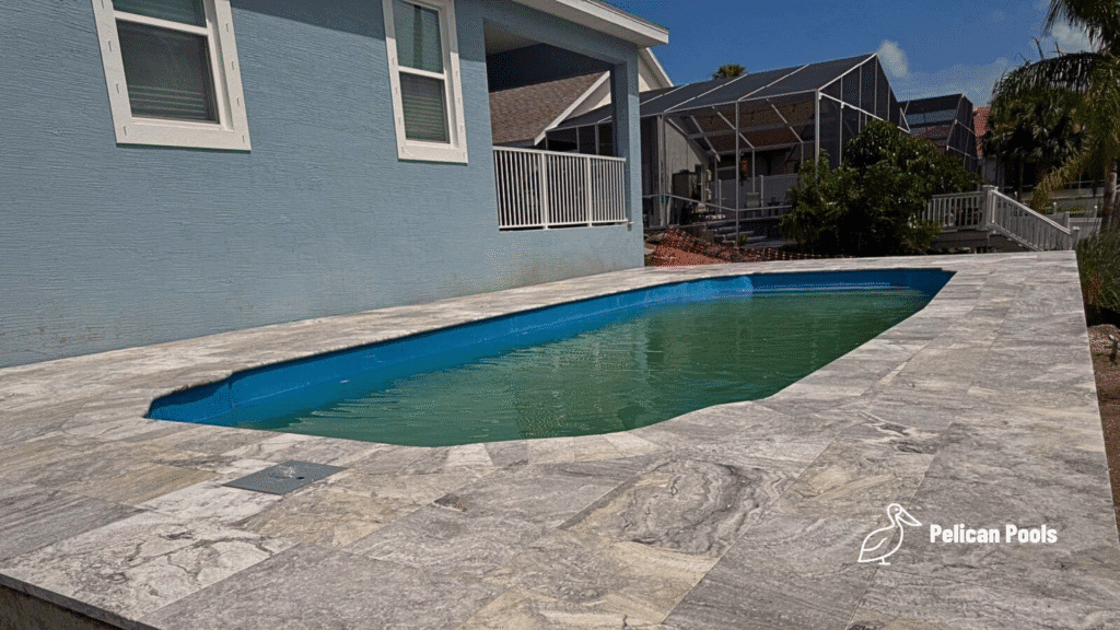 Fiberglass pool filled and surrounded by travertine deck next to home.