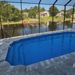 Blue fiberglass pool with travertine deck enclosed by screen overlooking canal.