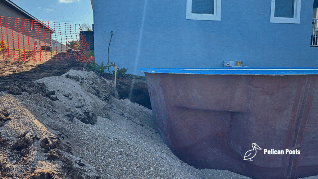 Fiberglass pool shell positioned close to house wall, surrounded by gravel.