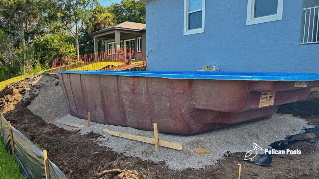 Fiberglass pool shell partially installed beside blue house.