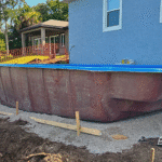 Fiberglass pool shell partially installed beside blue house.