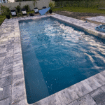 Rectangular blue-water fiberglass pool with gray paver coping and poolside lounge chairs.