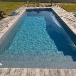 Rectangular blue-water fiberglass pool with gray paver coping and poolside lounge chairs.