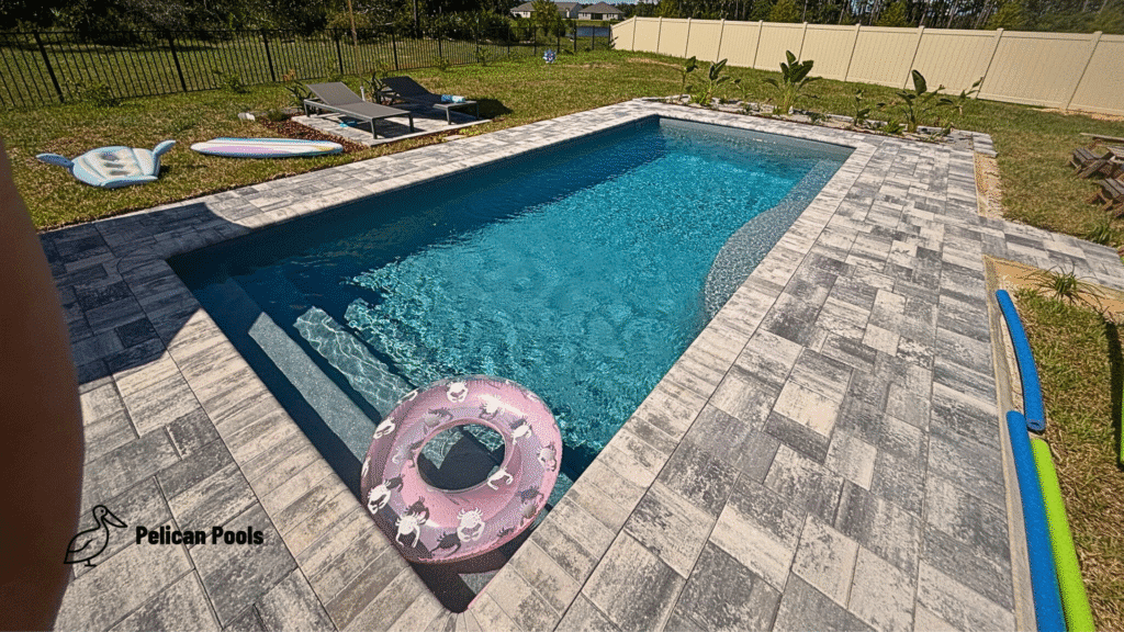 Pool with gray paver deck, sun loungers beside a grass lawn and garden plantings.