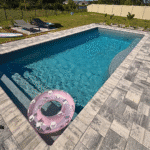 Pool with gray paver deck, sun loungers beside a grass lawn and garden plantings.
