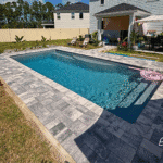 Modern fiberglass pool surrounded by gray paver deck, backyard home in background.