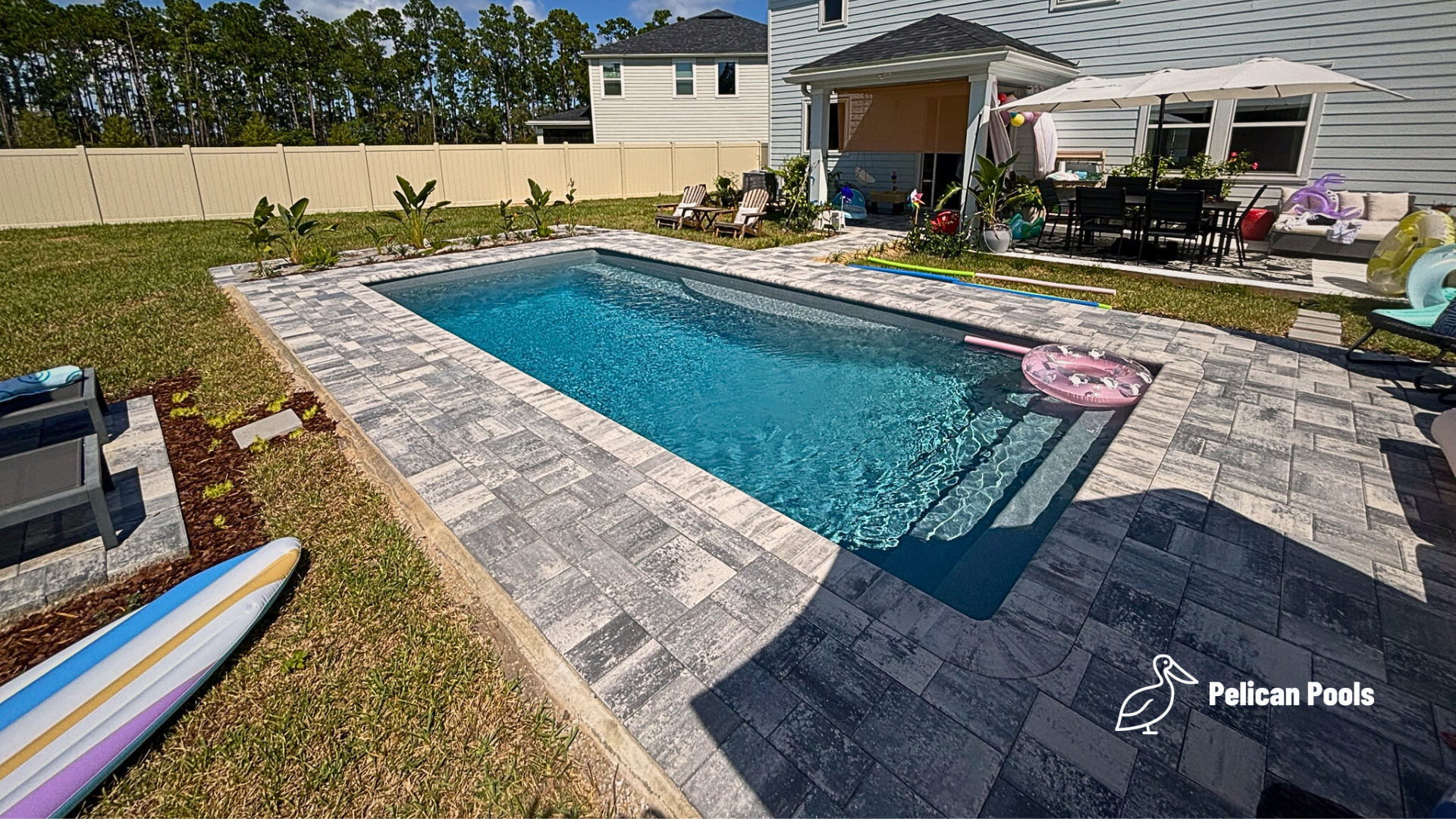 Modern fiberglass pool surrounded by gray paver deck, backyard home in background.