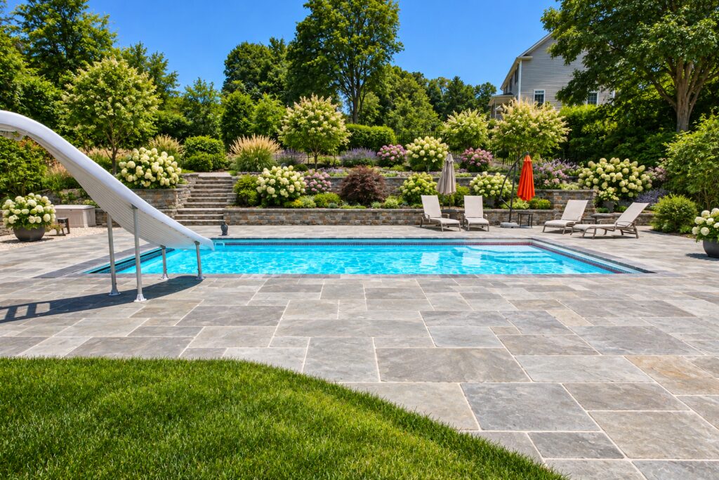 Side view of a San Juan Monte Carlo pool with a slide, gray natural stone paving and terraced hydrangea beds